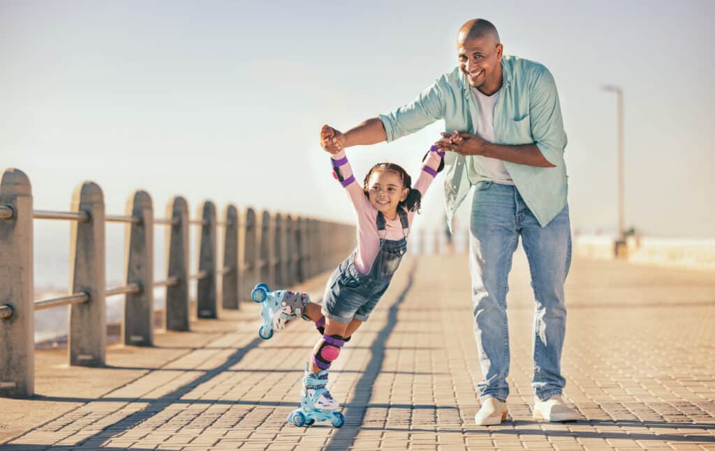 Father holding his child’s hands while she learns to roller skate on a beach promenade, a warm moment that reflects how the child tax credit supports everyday family life.