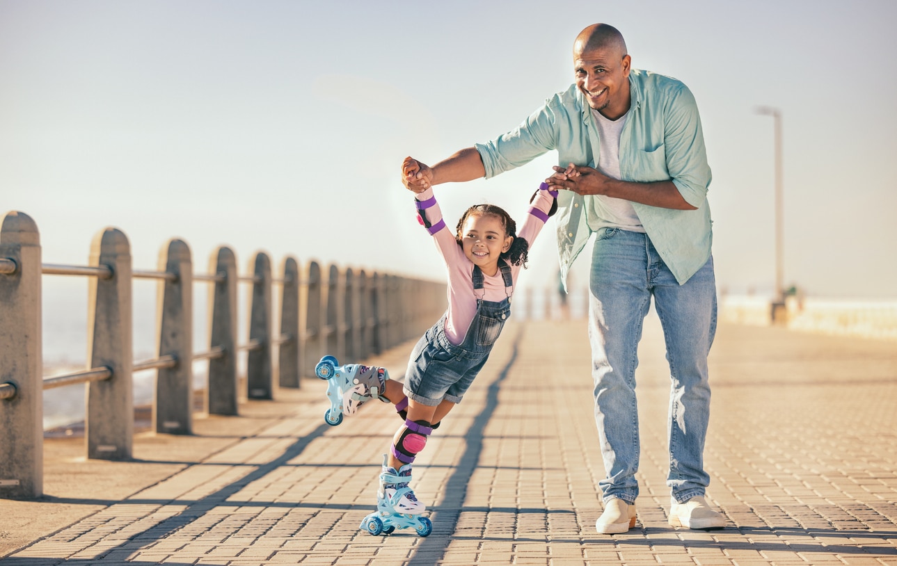 Father holding his child’s hands while she learns to roller skate on a beach promenade, a warm moment that reflects how the child tax credit supports everyday family life.