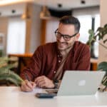 Young businessman working on a laptop and writing notes, planning around international payroll issues like a totalization agreement.