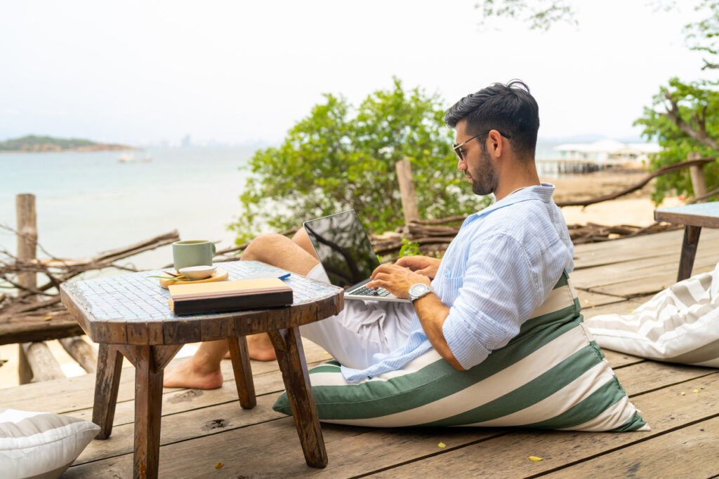 Caucasian businessman working remotely on a laptop at a beach café, managing digital nomad taxes while traveling.