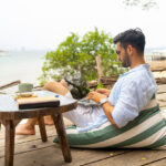 Caucasian businessman working remotely on a laptop at a beach café, managing digital nomad taxes while traveling.