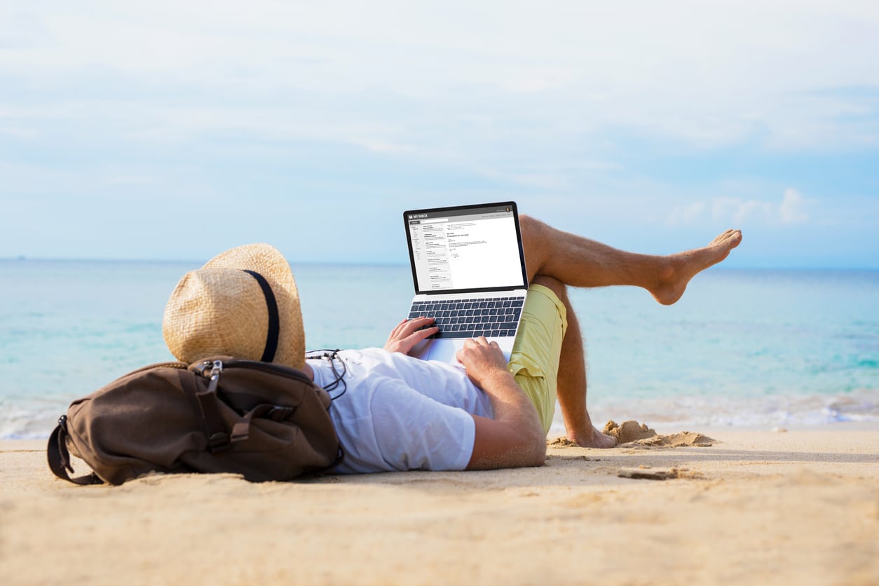 Man reading email on a laptop while relaxing on the beach, reflecting the self-employed lifestyle tied to Schedule SE.
