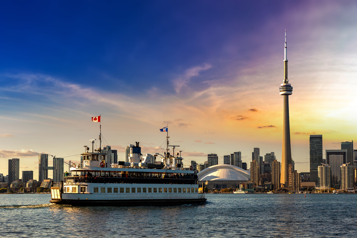 Panoramic view of the Toronto skyline and CN Tower at sunset, with a ferry crossing the water—showing city life and the cost of living in Canada.