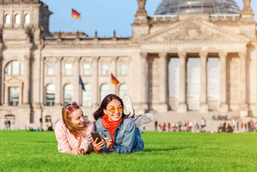 Two young women relaxing on the grass in front of the Bundestag in Berlin, enjoying student life while immersing themselves in the diverse languages spoken in Germany.