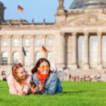 Two young women relaxing on the grass in front of the Bundestag in Berlin, enjoying student life while immersing themselves in the diverse languages spoken in Germany.